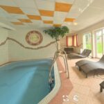 Innenpoolbereich mit kleinem Schwimmbecken, gefliestem Boden, mehreren Korbsesseln, Topfbaum, Wandkunst und großen Fenstern, die viel natürliches Licht hereinlassen.