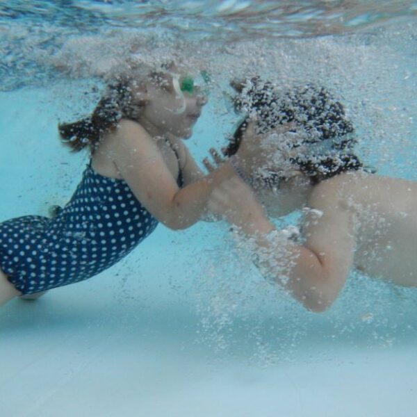 Zwei Kinder in Badeanzügen und mit Schwimmbrillen schwimmen in einem Becken unter Wasser, einander zugewandt und mit Luftblasen um ihre Gesichter.