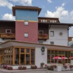 Ein mehrstöckiges Gästehaus mit Balkonen, Blumenkästen und Sitzgelegenheiten im Freien mit roten Sonnenschirmen vor der Kulisse von Bergen und Bäumen.