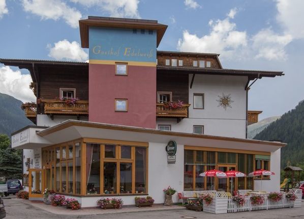 Ein mehrstöckiges Gästehaus mit Balkonen, Blumenkästen und Sitzgelegenheiten im Freien mit roten Sonnenschirmen vor der Kulisse von Bergen und Bäumen.