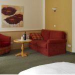 Hotelzimmer mit einem Doppelbett, einem roten Sessel und einem Sofa, einem kleinen runden Tisch mit Flaschen, einem Fernseher auf einer Kommode, Wandkunst und einer offenen Tür, die zu einem Badezimmer führt.