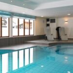 Innenschwimmbad mit klarem, blauem Wasser, umgeben von Liegestühlen und großen Fenstern, die natürliches Licht hereinlassen.