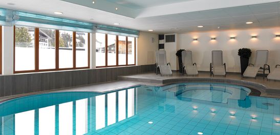 Innenschwimmbad mit klarem, blauem Wasser, umgeben von Liegestühlen und großen Fenstern, die natürliches Licht hereinlassen.