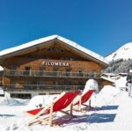 Ein hölzernes Hotel namens Filomena steht in einer verschneiten Berglandschaft mit zwei roten Liegestühlen davor und Menschen, die in der Nähe unter einem klaren blauen Himmel spazieren gehen.