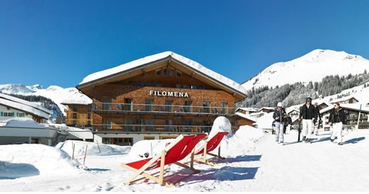 Ein hölzernes Hotel namens Filomena steht in einer verschneiten Berglandschaft mit zwei roten Liegestühlen davor und Menschen, die in der Nähe unter einem klaren blauen Himmel spazieren gehen.
