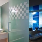 Modernes Spa-Interieur mit einer Trennwand aus Milchglas, einem Waschbecken mit einem Naturwandbild und einem Entspannungsbereich mit geschwungenen Liegestühlen unter blauer Beleuchtung.