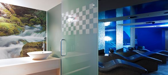 Modernes Spa-Interieur mit einer Trennwand aus Milchglas, einem Waschbecken mit einem Naturwandbild und einem Entspannungsbereich mit geschwungenen Liegestühlen unter blauer Beleuchtung.