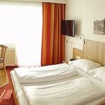 Ein Hotelzimmer mit einem Doppelbett, einem hölzernen Kopfteil, einem an der Wand montierten Fernseher, einem kleinen Tisch mit zwei Stühlen, einem roten Teppich und gestreiften Vorhängen vor einem großen Fenster.