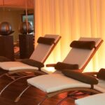 Drei moderne Loungesessel mit weißen Polstern und braunen Kissen sind vor einem sanft beleuchteten, beigen Vorhang aufgereiht. Zwei dekorative Kugeln stehen auf Ständern im Hintergrund.