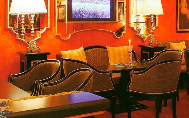 Eine gemütliche Sitzecke mit gepolsterten Stühlen, kleinen Tischen, beleuchteten Lampen und einem Fernseher an einer rot-orangen Wand.