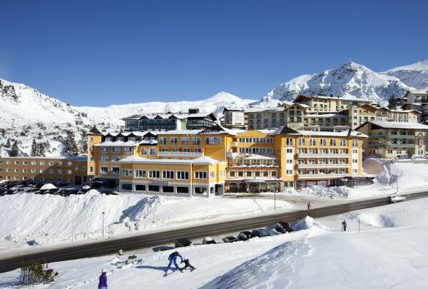 Ein großer gelber Hotelkomplex liegt auf einem verschneiten Hügel mit Bergen im Hintergrund; Menschen gehen in der Nähe des Gebäudes spazieren und Autos sind entlang der Straße geparkt.