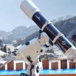 Im Freien ist ein Teleskop auf einem Stativ montiert, im Hintergrund sind schneebedeckte Berge, Bäume und ein klarer Himmel zu sehen.