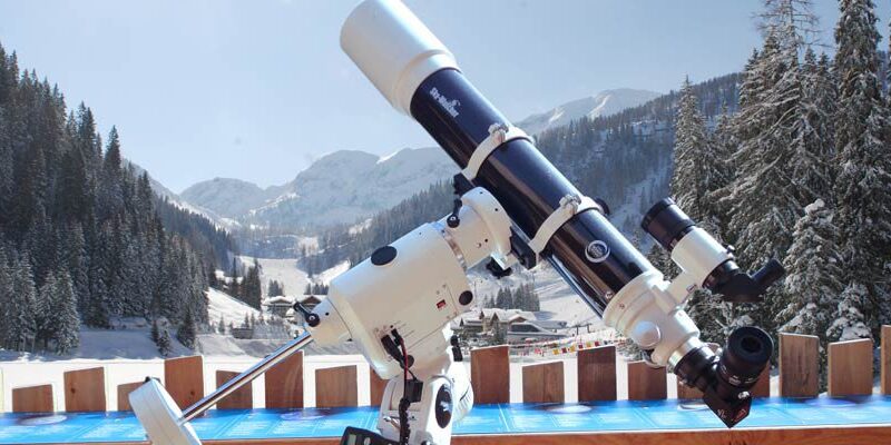 Im Freien ist ein Teleskop auf einem Stativ montiert, im Hintergrund sind schneebedeckte Berge, Bäume und ein klarer Himmel zu sehen.