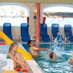 Mehrere Kinder und Erwachsene spielen in einem Hallenbad neben einer grün-gelben Rutsche, während im Hintergrund blaue Liegestühle aufgereiht sind.
