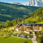 Ein großes Alpenhotel mit Sonnenkollektoren liegt in einem grünen Tal, umgeben von bewaldeten Hügeln und fernen Bergen, unter einem klaren blauen Himmel.