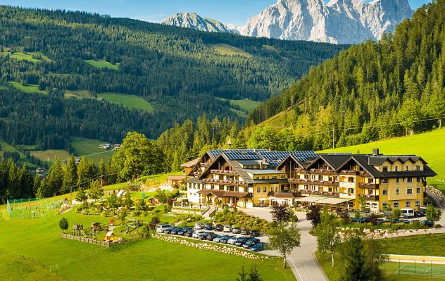 Ein großes Alpenhotel mit Sonnenkollektoren liegt in einem grünen Tal, umgeben von bewaldeten Hügeln und fernen Bergen, unter einem klaren blauen Himmel.