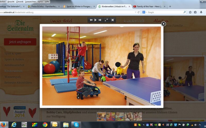 Kinder spielen auf Matten und fahren mit Spielzeug in einem Indoor-Spielbereich, während eine Frau neben einer blauen Tischtennisplatte im Vordergrund steht.