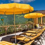 Eine Terrasse mit gestreiften gelben Liegestühlen und passenden gelben Sonnenschirmen bietet einen Blick auf eine malerische Berglandschaft unter blauem Himmel.