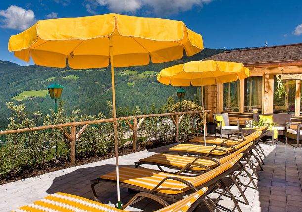 Eine Terrasse mit gestreiften gelben Liegestühlen und passenden gelben Sonnenschirmen bietet einen Blick auf eine malerische Berglandschaft unter blauem Himmel.