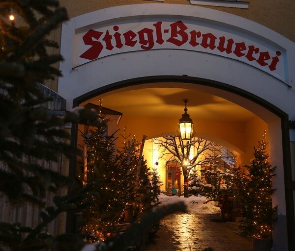 Der Eingang zur Stiegl-Brauerei ist mit Weihnachtsbäumen und Lichtern geschmückt, im Torbogen hängt eine Laterne, im Hintergrund ist Schnee zu sehen.