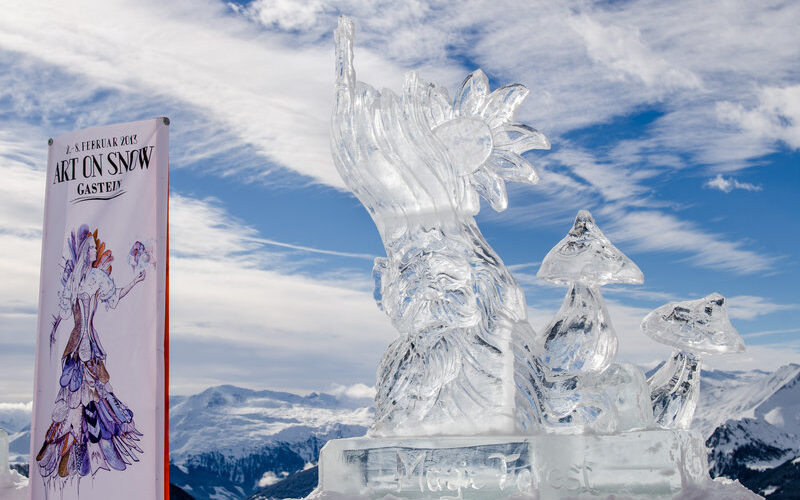 Eine klare Eisskulptur einer bärtigen Figur mit einem hohen Hut und zwei Pilzen wird im Freien auf Schnee ausgestellt, mit Bergen und einem „Art on Snow“-Veranstaltungsbanner im Hintergrund.