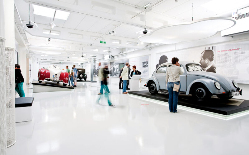 Menschen betrachten Oldtimer in einer hellen, modernen Ausstellung im Automobilmuseum mit weißen Böden und Wänden.