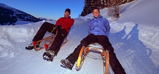 Zwei Personen in Winterjacken und Stiefeln sitzen auf Holzschlitten auf einem schneebedeckten Hang, umgeben von schneebedeckten Bäumen und Bergen unter einem klaren blauen Himmel.