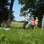 Mehrere Kinder stehen auf einer Wiese im Freien, während ein Junge nach vorne springt. Ein Erwachsener sitzt in der Nähe auf dem Boden und beobachtet. Im Hintergrund sind Bäume und Felder zu sehen.