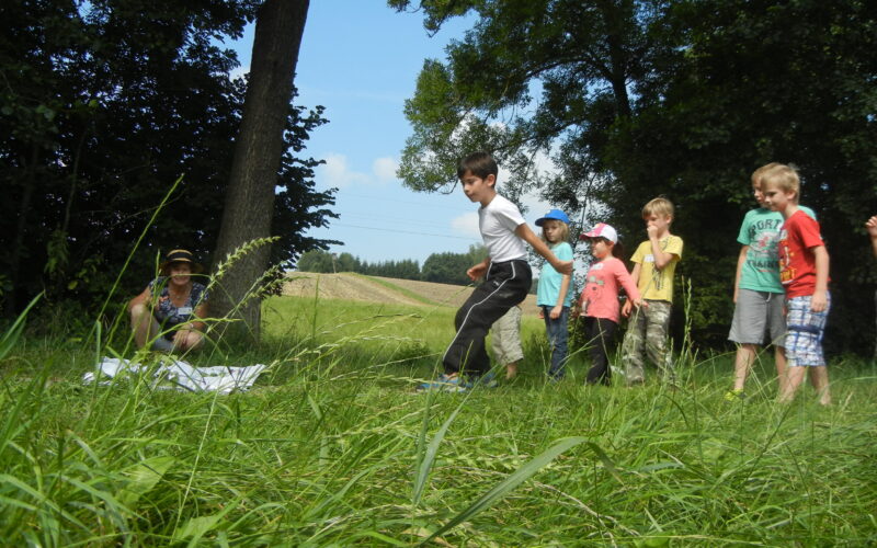 Mehrere Kinder stehen auf einer Wiese im Freien, während ein Junge nach vorne springt. Ein Erwachsener sitzt in der Nähe auf dem Boden und beobachtet. Im Hintergrund sind Bäume und Felder zu sehen.