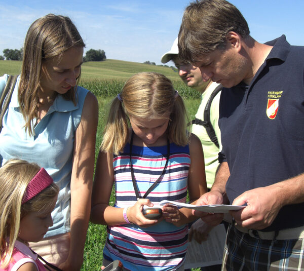 Vier Personen und ein Kind stehen an einem sonnigen Tag draußen auf einer Wiese und schauen auf eine Karte und ein GPS-Gerät.