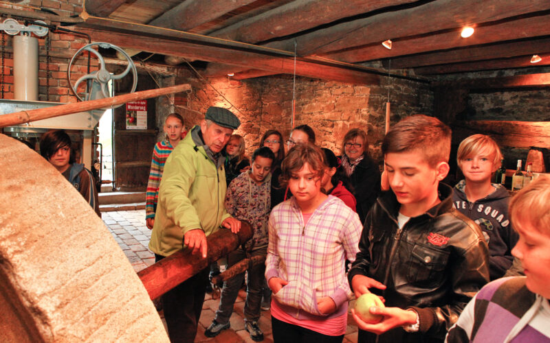 Eine Gruppe von Kindern und ein Erwachsener stehen in einem rustikalen Gebäude mit Backsteinwänden und Holzbalken und betrachten alte Maschinen.