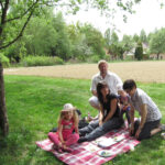Eine fünfköpfige Familie sitzt auf einer Decke und macht ein Picknick unter einem Baum in der Nähe eines Feldes. Auf der Decke stehen Teller mit Essen, im Hintergrund sind Häuser zu sehen.