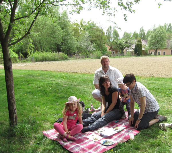 Eine fünfköpfige Familie sitzt auf einer Decke und macht ein Picknick unter einem Baum in der Nähe eines Feldes. Auf der Decke stehen Teller mit Essen, im Hintergrund sind Häuser zu sehen.