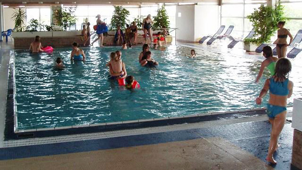 Mehrere Personen, darunter Kinder und Erwachsene, schwimmen und spielen in einem Hallenbad; einige stehen oder sitzen am Beckenrand.