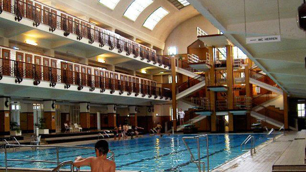 Hallenbad mit mehreren Sprungtürmen, Balkonen und Menschen, die schwimmen oder am Wasser sitzen. Das Sonnenlicht fällt durch gewölbte Fenster von oben ein.