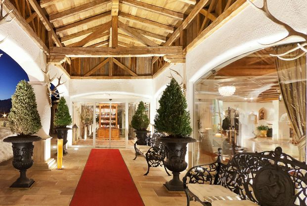 Überdachter Eingang mit Holzbalken, rotem Teppich, Topiary-Pflanzen, schwarzen, verzierten Bänken und Glastüren, die in einen hellen Innenraum führen.