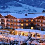 Ein großes Alpenhotel mit leuchtenden Lichtern steht in einer verschneiten Berglandschaft in der Abenddämmerung, umgeben von schneebedeckten Bäumen und Hügeln.