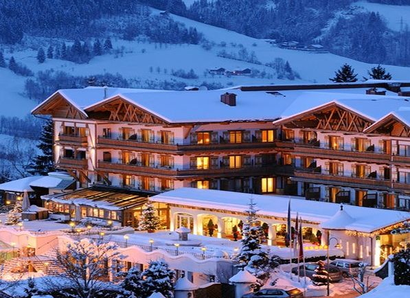 Ein großes Alpenhotel mit leuchtenden Lichtern steht in einer verschneiten Berglandschaft in der Abenddämmerung, umgeben von schneebedeckten Bäumen und Hügeln.