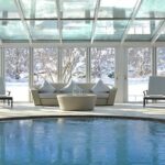 Sitzbereich im Innenbereich mit rundem Tisch und Stühlen neben dem Pool, umgeben von Glaswänden, die eine verschneite Landschaft nach draußen zeigen.