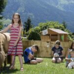 Fünf Kinder interagieren mit einem Pony und Kaninchen auf einer Wiese, mit einer kleinen Holzhütte und Bergen im Hintergrund.