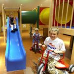 Drei Kinder spielen drinnen; eines rutscht eine blaue Rutsche hinunter, und zwei andere fahren mit Dreirädern in der Nähe eines hölzernen Spielgerüsts mit einem Tunnel.