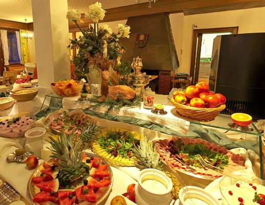 Ein Buffet mit verschiedenen Obstsorten, Salaten, Aufschnitt und Desserts, dekorativen Blumen und einem Korb mit Äpfeln steht in einem gut beleuchteten Essbereich.