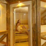 Eine Frau mit einem Handtuch sitzt in einer Holzsauna mit Glasfenstern und rustikaler Holzverkleidung.