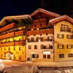 Ein warm beleuchtetes Hotel im alpinen Stil ist nachts von Schnee umgeben, mit Balkonen, Holzbalken und verschneiten Bäumen im Vordergrund.