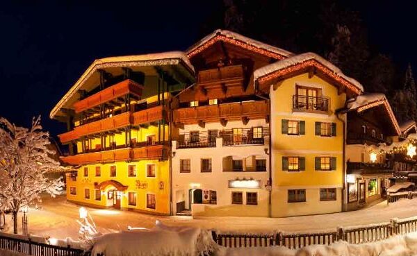 Ein warm beleuchtetes Hotel im alpinen Stil ist nachts von Schnee umgeben, mit Balkonen, Holzbalken und verschneiten Bäumen im Vordergrund.