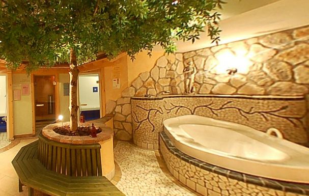 Wellnessbereich im Innenbereich mit einem großen Whirlpool aus Stein, Mosaikfliesenwänden, einem Baum in der Mitte mit einer runden Bank und warmer Beleuchtung.