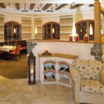 Ein gemütliches Restaurant mit Steinböden, gemusterten Sesseln, Weinregalen, Kerzen, Holzbalken und Tischen, an denen man in der Nähe von Bogenfenstern speisen kann.