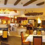 Ein hell erleuchtetes Restaurant mit gelben und orangefarbenen Stühlen, gedeckten Tischen, einem zentralen Kamin und hölzernen Deckenbalken.