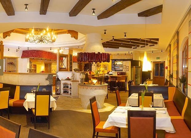 Ein hell erleuchtetes Restaurant mit gelben und orangefarbenen Stühlen, gedeckten Tischen, einem zentralen Kamin und hölzernen Deckenbalken.