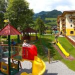 Kinder spielen auf bunten Rutschen und Klettergerüsten auf einem Spielplatz neben einem großen gelben Gebäude, mit grasbewachsenen Hügeln und Bergen im Hintergrund.